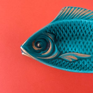 Könnte beinhalten: Eine türkisfarbene, fischförmige Keramikschale mit einer strukturierten Schuppenoptik auf dem Körper. Das Design weist goldfarbene Akzente auf, die das Auge, die Flossen und die Kiemendetails hervorheben und einen Kontrast zur türkisen Glasur bilden.