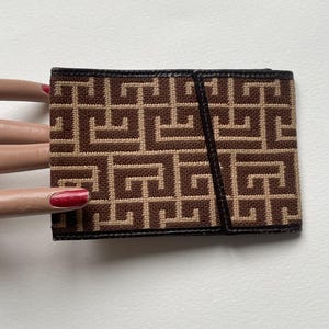 Puede incluir: Un tarjetero con estampado marrón y beige con un diseño geométrico. El tarjetero tiene un ribete de cuero negro y una correa de cuero negro. El diseño es un patrón repetitivo de formas entrelazadas.