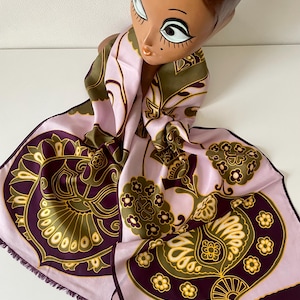 May include: A vintage silk scarf featuring a floral design in purple, olive green, and gold on a pale pink backdrop. The scarf, with a fringed edge, is draped over a decorative head.