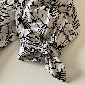 May include: Close-up of a white blouse sleeve with a black floral pattern. The sleeve is gathered at the wrist and tied with a bow. The fabric has a soft, flowing appearance.