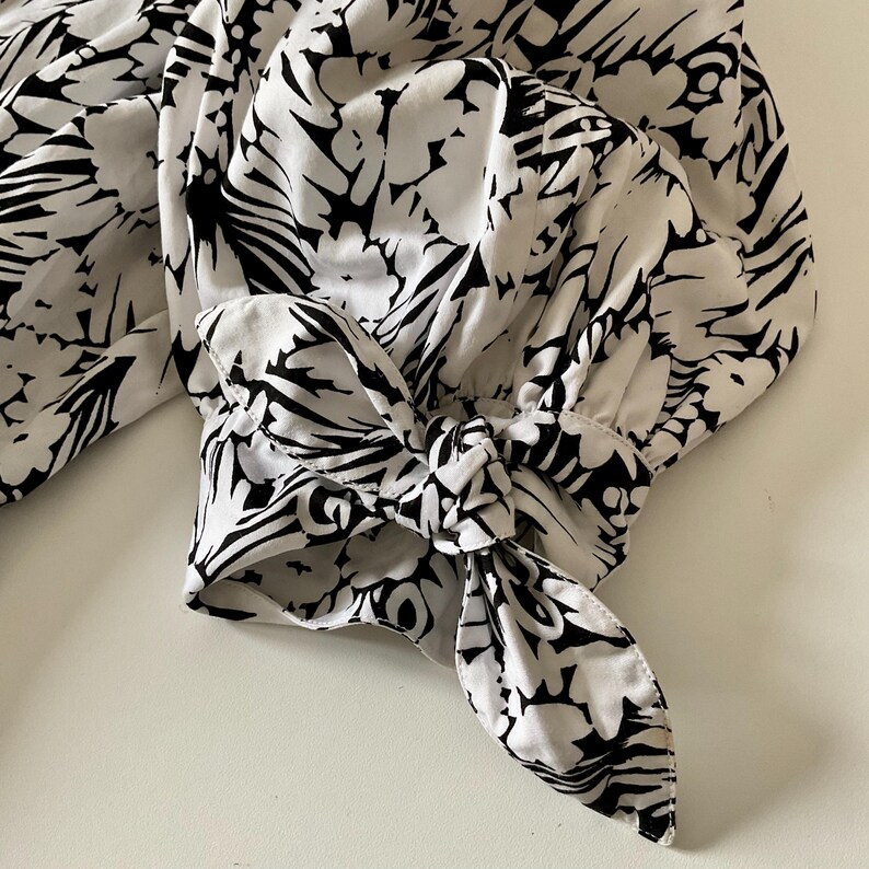 May include: Close-up of a white blouse sleeve with a black floral pattern. The sleeve is gathered at the wrist and tied with a bow. The fabric has a soft, flowing appearance.