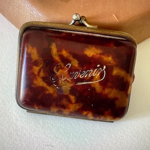 May include: A rectangular coin purse featuring a brown and amber tortoiseshell pattern with a metallic brass frame and a clasp closure. The word Souvenir is engraved in a cursive script across the center of the front surface of the purse.