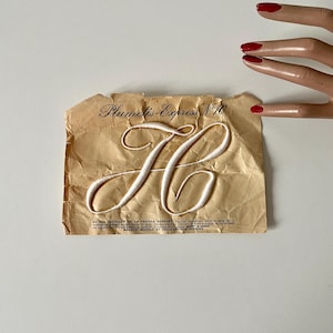Peut inclure: Un papier vintage avec le texte "Plumetis-Express N.10" et une lettre "H" brodée en blanc. Le papier est vieilli avec une texture froissée. Une main de mannequin avec du vernis à ongles rouge est dans le coin supérieur droit.