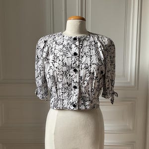 May include: A black and white floral print blouse with short sleeves and black buttons. The blouse is displayed on a mannequin. The sleeves are tied at the elbows. The blouse is a cropped style.