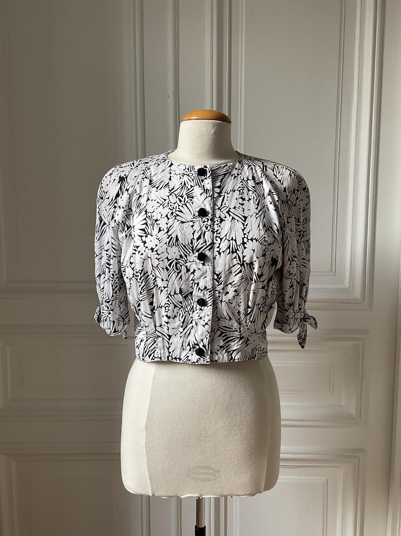 May include: A black and white floral print blouse with short sleeves and black buttons. The blouse is displayed on a mannequin. The sleeves are tied at the elbows. The blouse is a cropped style.