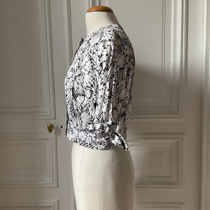 May include: A white and black floral print blouse with short sleeves and a tie detail at the waist. The blouse is buttoned up and displayed on a mannequin. The floral pattern is a mix of black and white, creating a striking contrast.