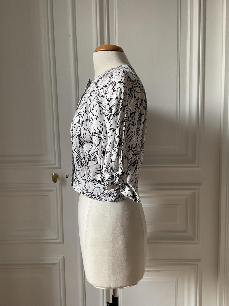 May include: A white and black floral print blouse with short sleeves and a tie detail at the waist. The blouse is buttoned up and displayed on a mannequin. The floral pattern is a mix of black and white, creating a striking contrast.