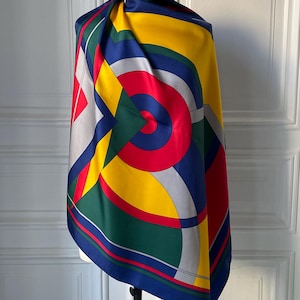 May include: A colorful silk scarf with a geometric design. The scarf features a vibrant pattern of red, yellow, blue, green, and gray shapes. The scarf is draped over a mannequin, showcasing its bold and eye-catching design.