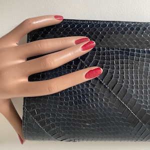 Peut inclure: Une pochette bleu marine foncé présentant un motif texturé en peau de serpent avec une finition brillante. Cet accessoire rectangulaire possède une fermeture à rabat structurée avec des coutures diagonales visibles sur la surface.