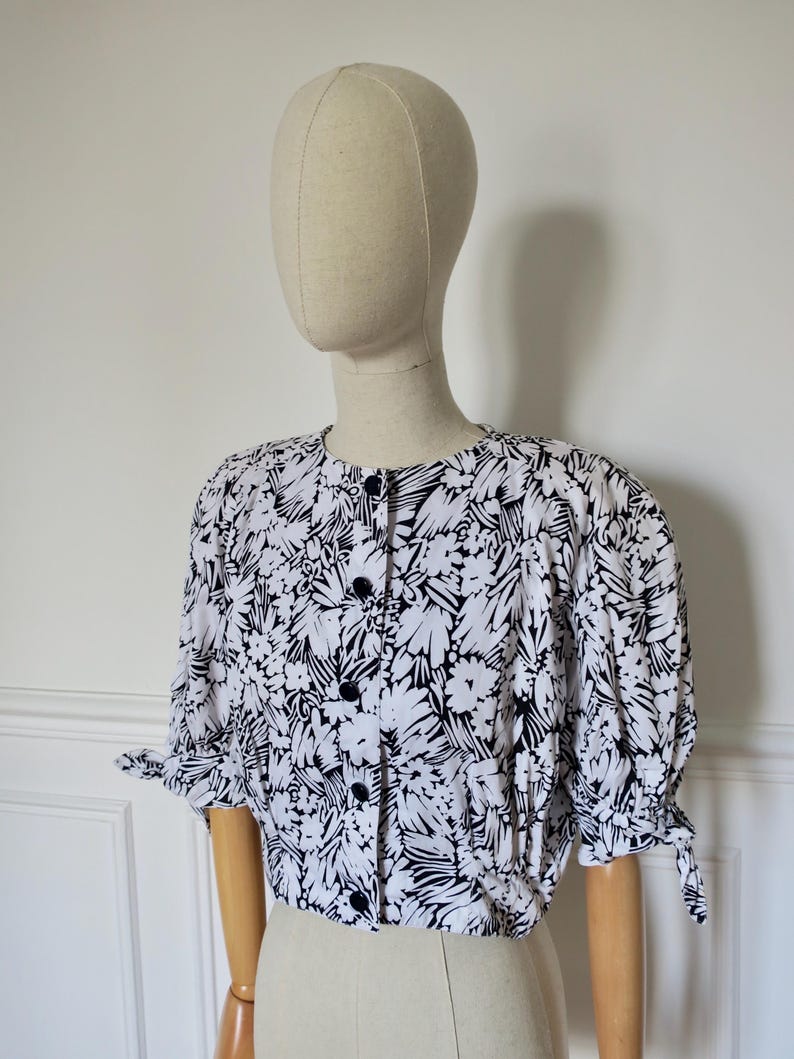 Peut inclure: Un chemisier à imprimé floral noir et blanc. Le tissu blanc est orné de motifs floraux noirs abstraits. Le chemisier a un col rond, des manches courtes froncées avec des détails de nœuds et une fermeture boutonnée avec des boutons noirs.