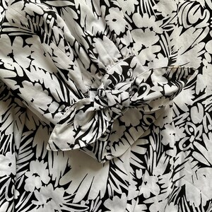 May include: Close-up of a white fabric with a bold black floral pattern. The sleeve cuff features a bow detail, also in the black and white floral print. The fabric appears to be lightweight, with soft folds and gathers.