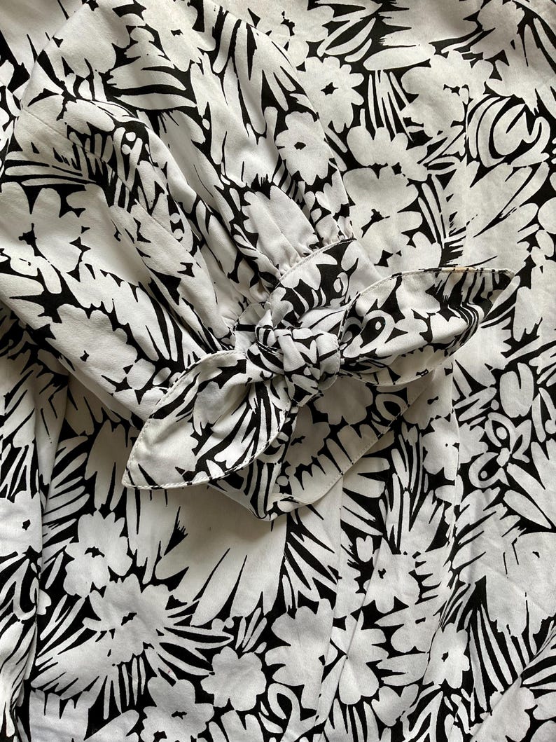 May include: Close-up of a white fabric with a bold black floral pattern. The sleeve cuff features a bow detail, also in the black and white floral print. The fabric appears to be lightweight, with soft folds and gathers.
