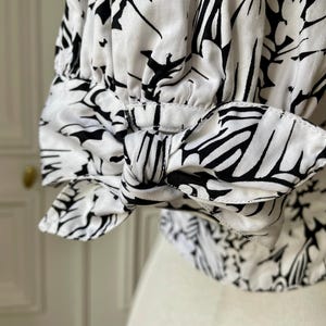 May include: Close-up of a white blouse sleeve with a black floral pattern. The sleeve features a bow detail at the cuff. The fabric is a light, flowing material, and the design is a bold, graphic print.