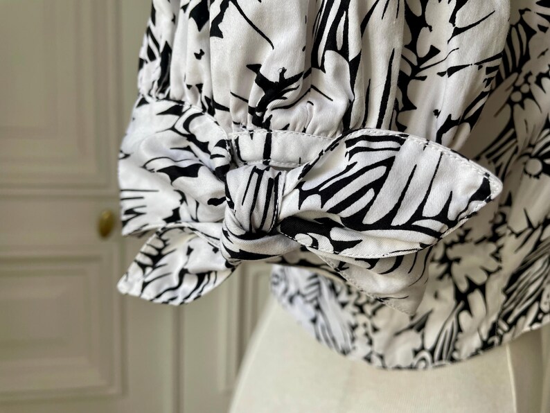 May include: Close-up of a white blouse sleeve with a black floral pattern. The sleeve features a bow detail at the cuff. The fabric is a light, flowing material, and the design is a bold, graphic print.