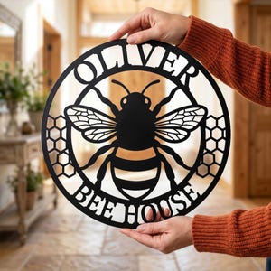 Könnte beinhalten: Schwarzes Metall-Bienenhaus-Schild mit dem Namen "OLIVER" über einer detaillierten Bienenillustration. Die Worte "BEE HOUSE" stehen darunter. Das Schild ist rund mit Waben-Details.