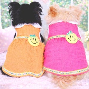 May include: Two dogs wearing matching dresses. One dog is wearing an orange dress, and the other is wearing a pink dress. Each dress has a white collar, a yellow smiley face, and a decorative trim.
