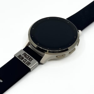 May include: A smartwatch with a round black face and a silver metallic casing. The watch features a black silicone band with a silver-toned metal keeper loop engraved with the text PACERS RUN CLUB in a bold, sans-serif font.