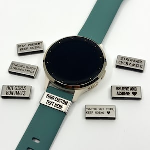 May include: A teal smartwatch with a round face and several rectangular metal charms featuring engraved text including STAY PRESENT KEEP GOING, STRONG BODY STRONG MIND, HOT GIRLS RUN HALFS, YOUR CUSTOM TEXT HERE, STRONGER EVERY MILE, BELIEVE AND ACHIEVE, and YOU'VE GOT THIS, KEEP GOING!