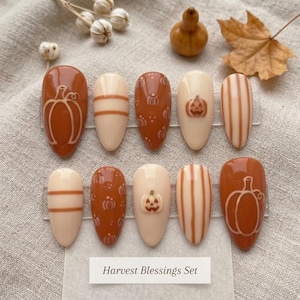 May include: A set of ten almond-shaped press-on nails featuring a fall-themed design in shades of burnt orange and cream. The nails display pumpkin illustrations, vertical stripes, and horizontal lines. A label reads Harvest Blessings Set.