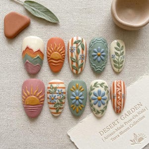 May include: A set of ten press-on nails featuring embossed designs of mountains, suns, flowers, and vines in earthy tones of terracotta, sage green, and cream. The text DESERT GARDEN | Artisan-Made Press-On Nails | Terra Bloom Collection is visible.