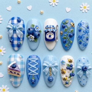 May include: A set of ten press-on nails featuring a blueberry theme with light blue and white colors. Designs include gingham patterns, 3D bows, blueberries, a jar labeled Blueberry Jam, bees, vines, a slice of cake, and lace-up corset details.