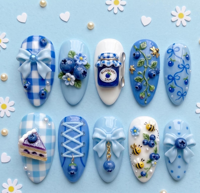 May include: A set of ten press-on nails featuring a blueberry theme with light blue and white colors. Designs include gingham patterns, 3D bows, blueberries, a jar labeled Blueberry Jam, bees, vines, a slice of cake, and lace-up corset details.
