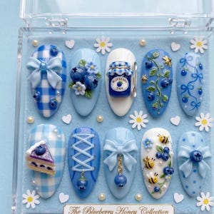 May include: A set of ten almond-shaped press-on nails in a clear case, featuring blue and white gingham, polka dots, blueberry motifs, 3D bows, a miniature jam jar labeled Blueberry Jam, cake slices, and bees on a light blue felt background.