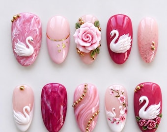 May include: A collection of ten oval-shaped artificial nails with various designs. The nails feature shades of pink, white, and gold. Some nails have swan and rose designs, while others have marble effects and gold accents.