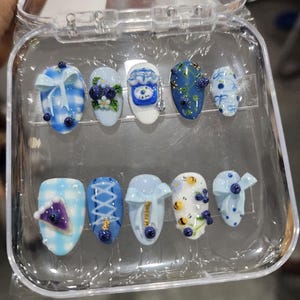 May include: A set of ten press-on nails in a clear plastic case, featuring various blue and white designs including gingham patterns, 3D bows, blueberries, small bees, a slice of purple pie, and intricate lace-up corset-style detailing on a glossy finish.