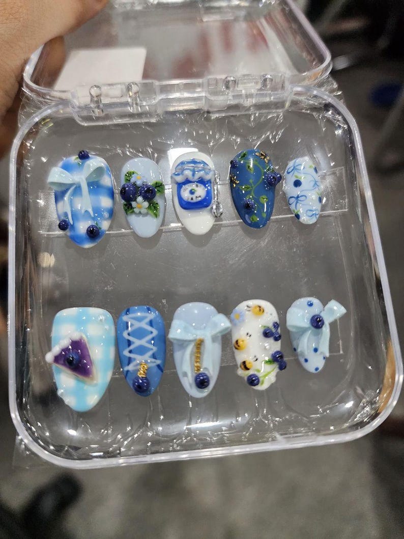 May include: A set of ten press-on nails in a clear plastic case, featuring various blue and white designs including gingham patterns, 3D bows, blueberries, small bees, a slice of purple pie, and intricate lace-up corset-style detailing on a glossy finish.