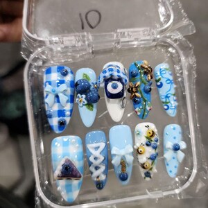 May include: A set of ten almond-shaped press-on nails in a clear plastic case. The designs feature light blue and white gingham patterns, 3D blue bows, painted blueberries, small bees, and a central eye motif. The number 10 is written on the lid.
