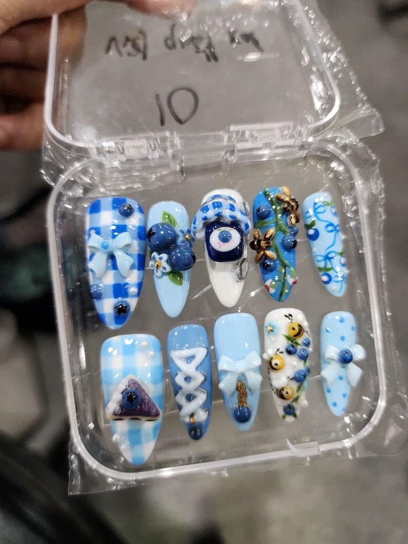 May include: A set of ten almond-shaped press-on nails in a clear plastic case. The designs feature light blue and white gingham patterns, 3D blue bows, painted blueberries, small bees, and a central eye motif. The number 10 is written on the lid.