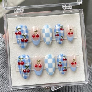 May include: A set of ten press-on nails in a clear plastic case. The nails feature a light blue and white checkered pattern, solid light blue tips, and light blue plaid designs with small, raised red cherry accents with green stems.