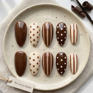 May include: A set of ten almond-shaped press-on nails featuring a brown and cream color palette. The designs include solid brown, cream with brown polka dots, brown with vertical cream stripes, brown with white polka dots, and cream with brown stripes.