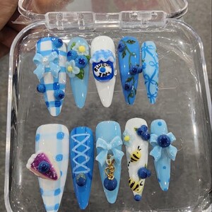May include: A set of ten almond-shaped press-on nails in a clear plastic case. The designs feature light blue and white color palettes with 3D accents including bows, blueberries, a jar labeled Blueberry Jam, a slice of pie, and small bumblebees.