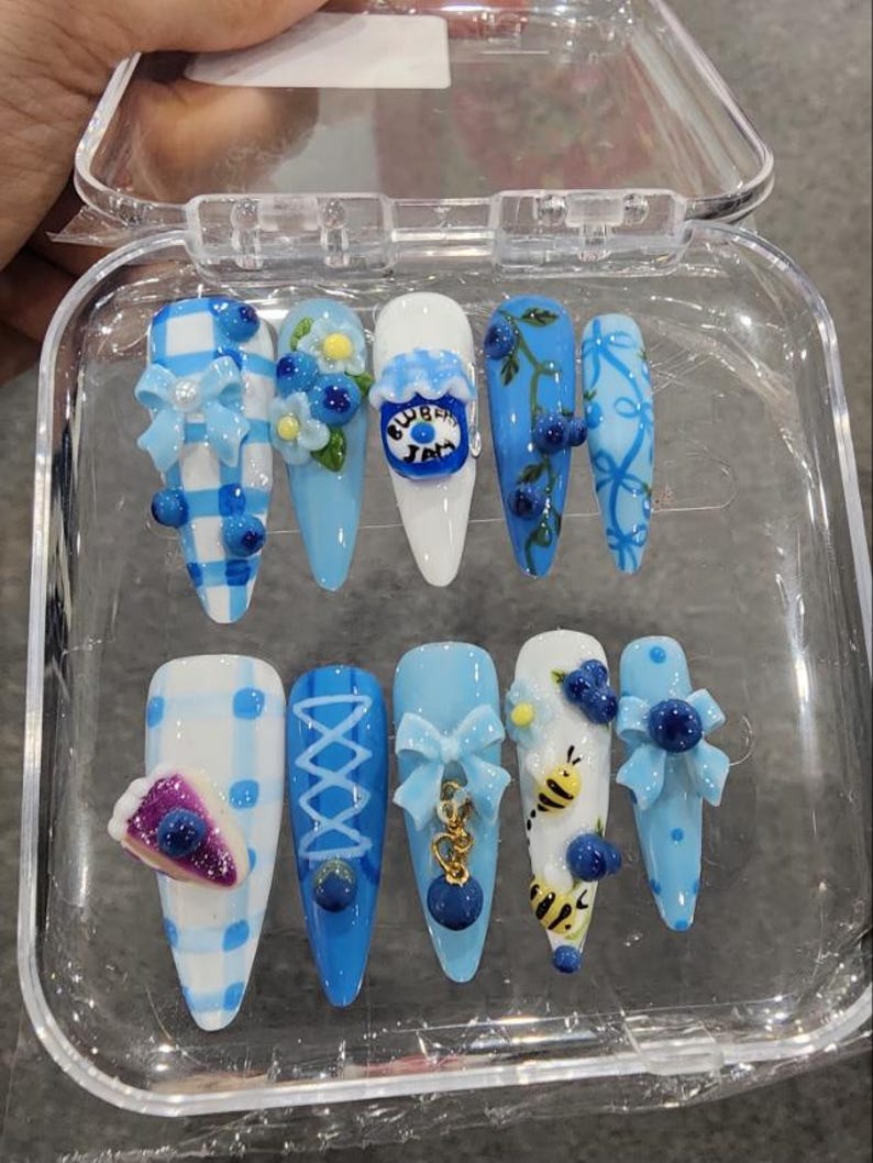 May include: A set of ten almond-shaped press-on nails in a clear plastic case. The designs feature light blue and white color palettes with 3D accents including bows, blueberries, a jar labeled Blueberry Jam, a slice of pie, and small bumblebees.