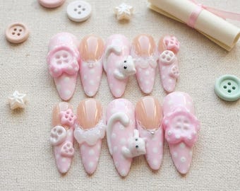 Puede incluir: Un conjunto de diez uñas artificiales rosas con lunares blancos y varios adornos 3D. Algunas uñas presentan un diseño de botón, otras un pequeño gato blanco y algunas tienen un ribete de encaje. Botones y decoraciones en forma de estrella están dispersos.