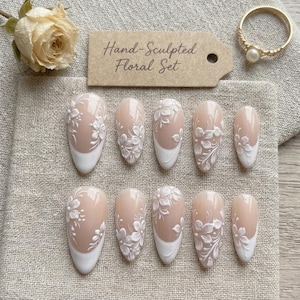May include: A set of ten almond-shaped press-on nails featuring a nude base with white 3D hand-sculpted floral designs and white French tips. A tan tag with the text Hand-Sculpted Floral Set is positioned above the nails, alongside a gold ring.