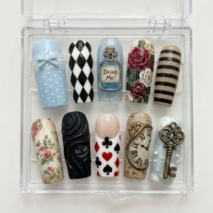 May include: A set of ten press-on nails in a clear plastic case featuring designs inspired by Alice in Wonderland, including a blue polka dot nail with a bow, black and white diamonds, a bottle labeled Drink Me!, roses, stripes, floral patterns, a black cat, playing card suits, a clock, and a key.