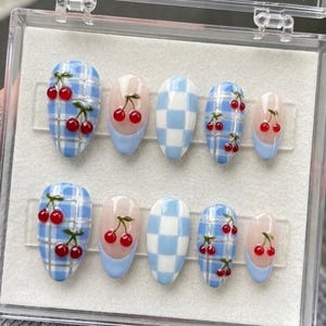 May include: A set of ten press-on nails in a clear plastic storage case. The designs include light blue and white checkered patterns, solid light blue French tips, and light blue plaid backgrounds decorated with small, glossy red cherry motifs.