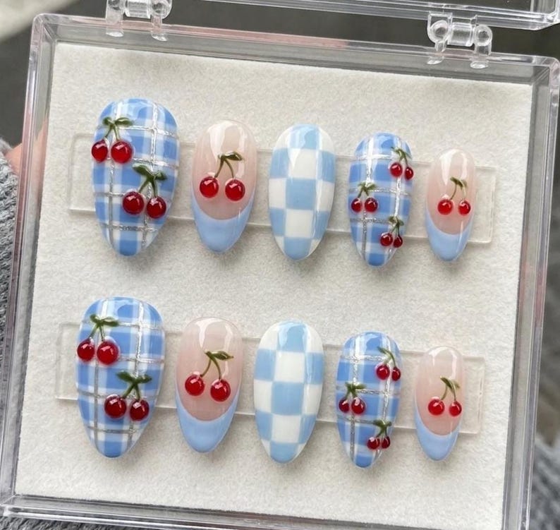 May include: A set of ten press-on nails in a clear plastic storage case. The designs include light blue and white checkered patterns, solid light blue French tips, and light blue plaid backgrounds decorated with small, glossy red cherry motifs.