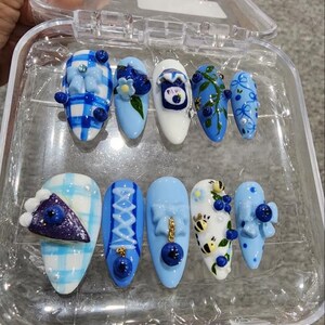 May include: A set of ten press-on nails in a clear plastic case, featuring light blue and white themes. Designs include blue gingham patterns, 3D blueberry accents, a miniature pie slice, small bees, bows, and a tiny jar illustration with blue details.