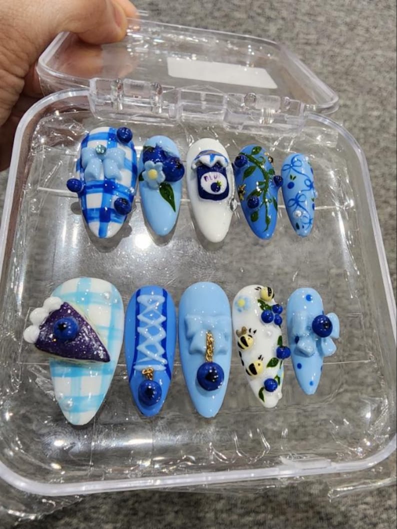May include: A set of ten press-on nails in a clear plastic case, featuring light blue and white themes. Designs include blue gingham patterns, 3D blueberry accents, a miniature pie slice, small bees, bows, and a tiny jar illustration with blue details.