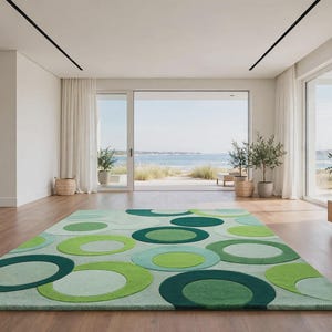 Op de afbeelding: Een rechthoekig vloerkleed met een herhalend patroon van overlappende cirkels in diverse groentinten, waaronder limoen, bosgroen en mint. Het kleed ligt op een lichte houten vloer in een kamer met witte muren en grote raampartijen.