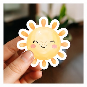 Puede incluir: Una pegatina troquelada con forma de sol amarillo con cara sonriente, ojos cerrados con pesta&ntilde;as, mejillas rosadas y pecas marrones. El sol tiene rayos redondeados y un borde blanco que rodea toda la figura.