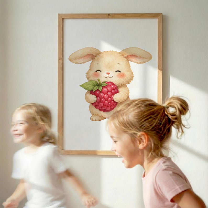 May include: A framed print featuring a watercolor illustration of a smiling, tan bunny holding a large red raspberry with green leaves. The artwork has a light wood frame and is displayed on a white wall. Two young girls are in the foreground.