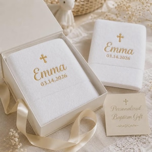 May include: Two white towels with gold embroidery featuring a cross, the name Emma, and the date 03.14.2026. One towel is inside an open white gift box with a ribbon, and the other is folded next to it with a card reading Personalized Baptism Gift.