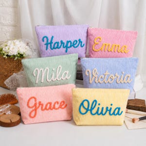 May include: Six rectangular corduroy pouches in various pastel colors, each featuring a name embroidered in a contrasting yarn script. The pouches are lavender with blue text, pink with yellow text, mint with white text, blue with beige text, peach with orange text, and yellow with blue text.