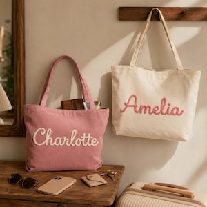 Könnte beinhalten: Zwei Cord-Einkaufstaschen mit zwei Schulterriemen. Eine Tasche ist altrosa mit dem in weißer Schreibschrift gestickten Namen Charlotte. Die andere Tasche ist cremefarben mit dem in rosa Schreibschrift gestickten Namen Amelia. Beide Taschen haben einen strukturierten Stoff.