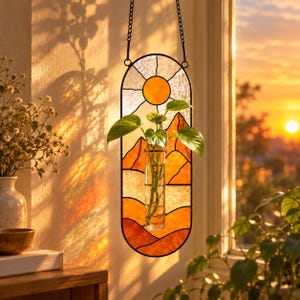 Könnte beinhalten: Ein vertikales, ovales Wandbild aus Buntglas mit einer Sonnen- und Berglandschaft in Orange- und Gelbtönen. In der Mitte ist ein durchsichtiges Glasreagenzglas befestigt, das einen grünen Pflanzensteckling mit mehreren Blättern enthält.